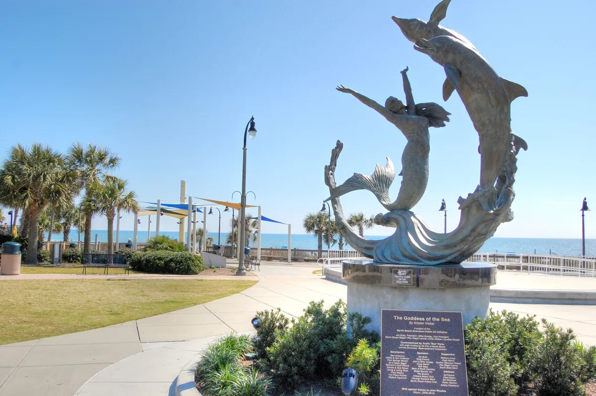 This exterior image features the iconic statue of the Goddess of the Sea along a scenic walking path lined with palm trees, with the ocean visible in the background. People are seen strolling, taking photos, and enjoying the tranquil beachside setting.