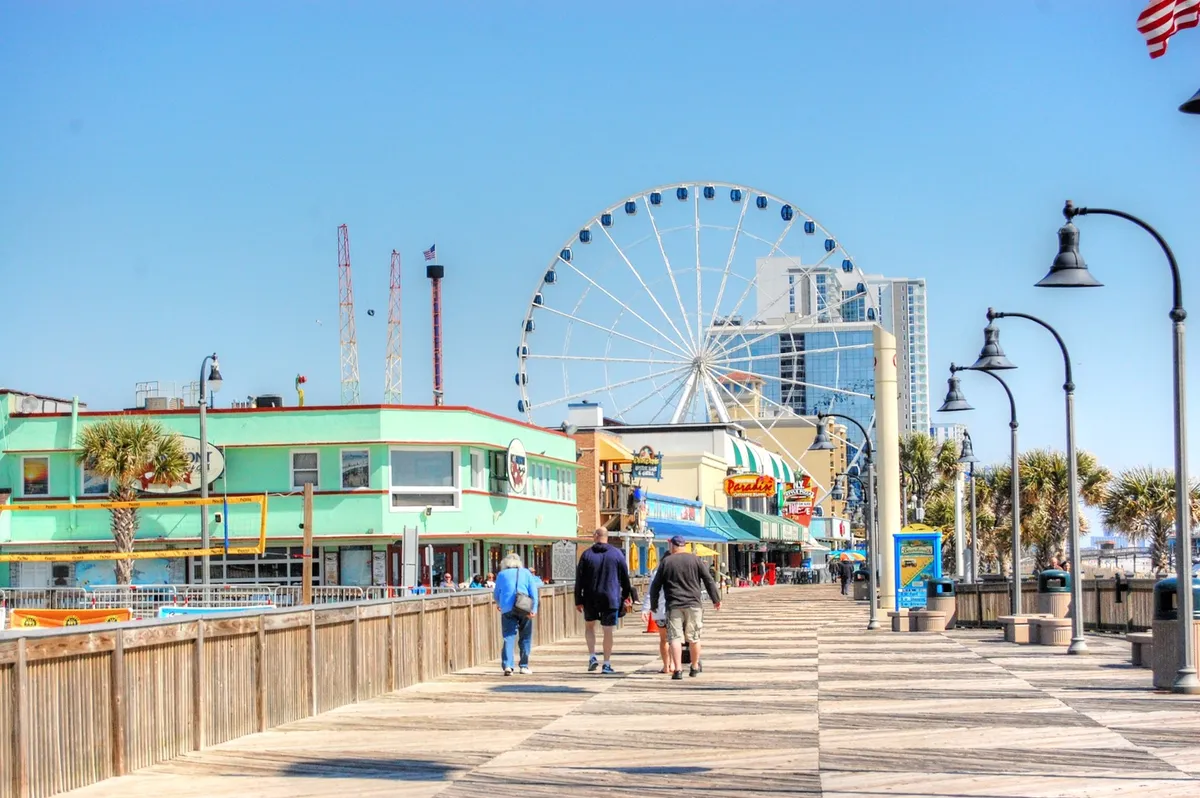 An engaging boardwalk scene featuring the Ferris wheel (another term often used for SkyWheel), colorful storefronts, bustling crowds, and lush palm trees. The joyful outdoor ambiance highlights Myrtle Beach’s lively attractions, shopping, and dining experiences available to visitors.