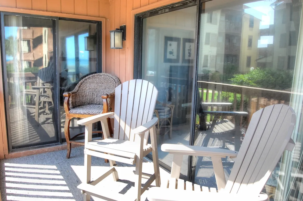 A serene balcony with comfortable Adirondack and woven chairs arranged near glass sliding doors. The setup invites quiet relaxation, reading, or enjoying the ocean breeze, and the vantage point reinforces the indoor-outdoor connection.