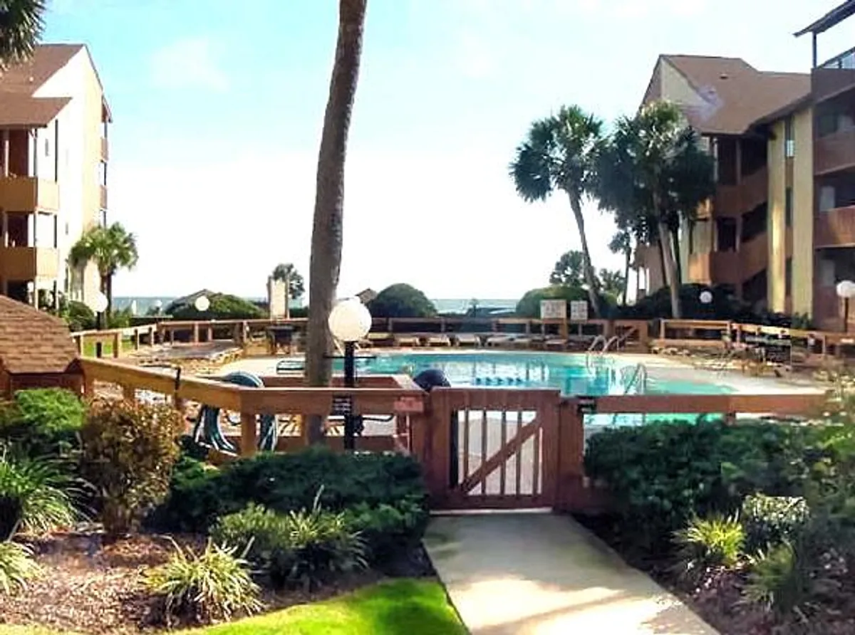 A wide-angle perspective of the outdoor swimming pool area, framed by lush palm trees and nearby buildings. The gated entryway hints at privacy and security, while the open space is ideal for family gatherings or community activities.