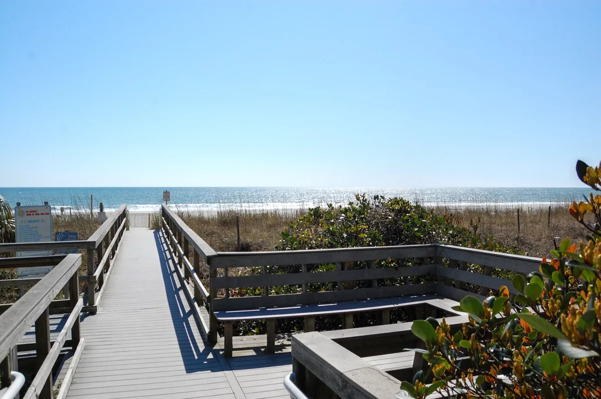 A wide wooden walkway leads through beach grasses toward a pristine stretch of ocean. The scene includes the open beach, gently rolling waves, and a soft horizon, all bathed in natural light. The setting is peaceful and inviting, perfect for illustrating direct access to the uncrowded sands.