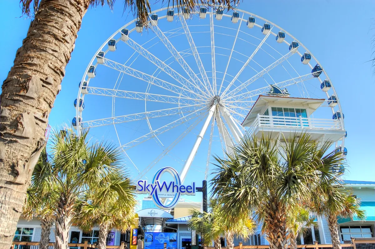 This vibrant exterior image features the iconic Myrtle Beach SkyWheel, framed by swaying palm trees and the entry area bustling with visitors. The joyful mood and panoramic perspective convey the thrill and excitement of one of Myrtle Beach’s most recognizable attractions.