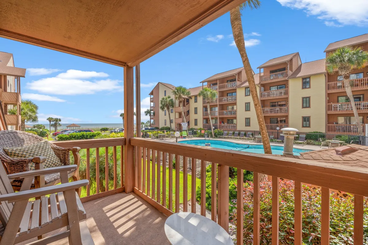 An exterior balcony scene with comfortable seating overlooking the pool, palm trees, and neighboring buildings. The balcony invites guests to relax, dine outdoors, and soak in the ocean-inspired atmosphere, hinting at ocean views and breezes.