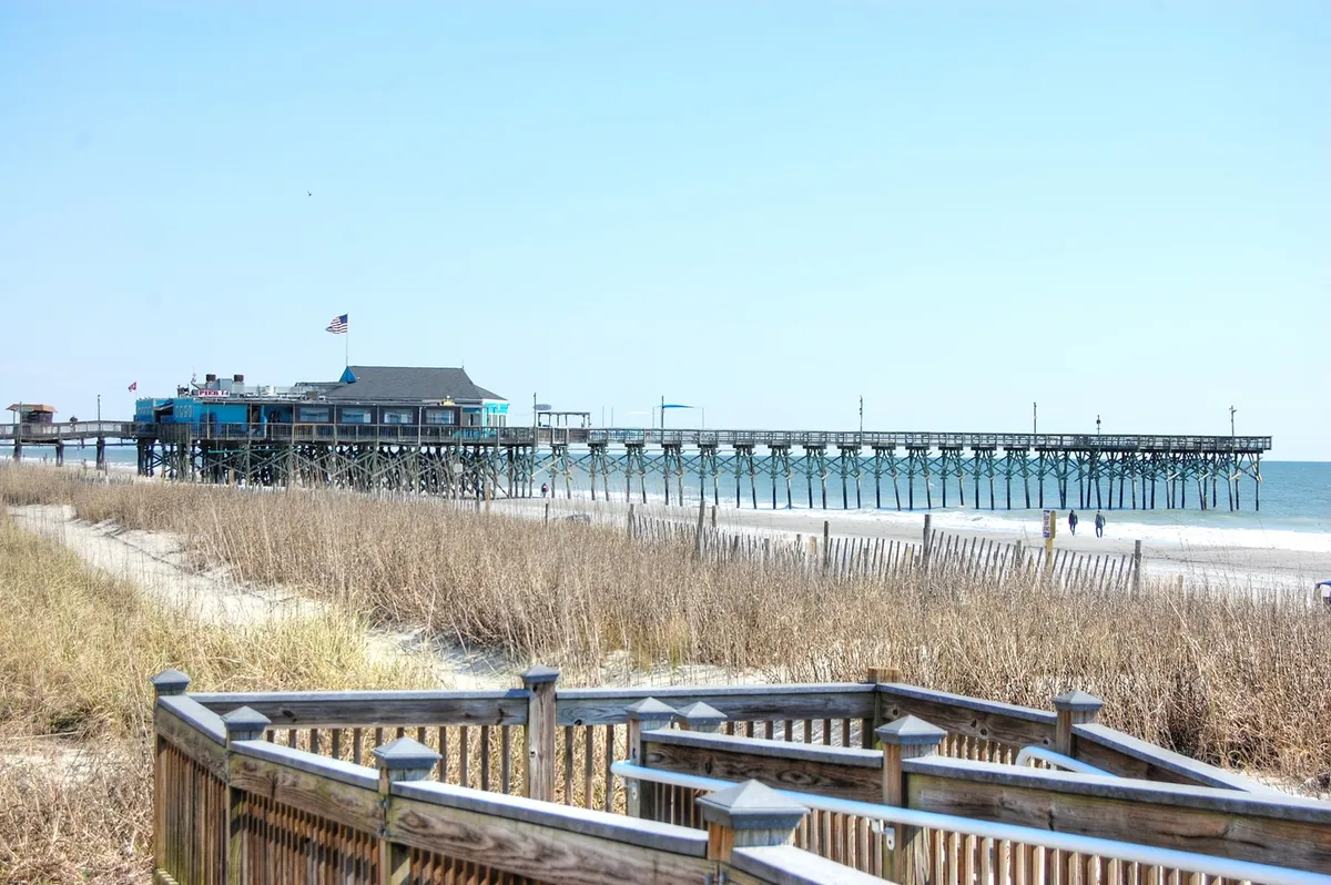 A tranquil scene of a pier stretching out into the sea, bordered by sandy beach and grass. The beach offers inviting space for sunbathing, sandcastle building, and leisurely walks, with the vast ocean creating a serene coastal backdrop.