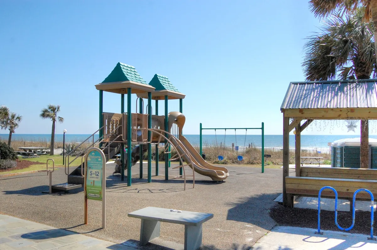 A sunny seaside playground scene set against the backdrop of ocean views and palm trees. Families and children are enjoying playground equipment and swinging, illustrating carefree, outdoor fun along the Myrtle Beach coast.