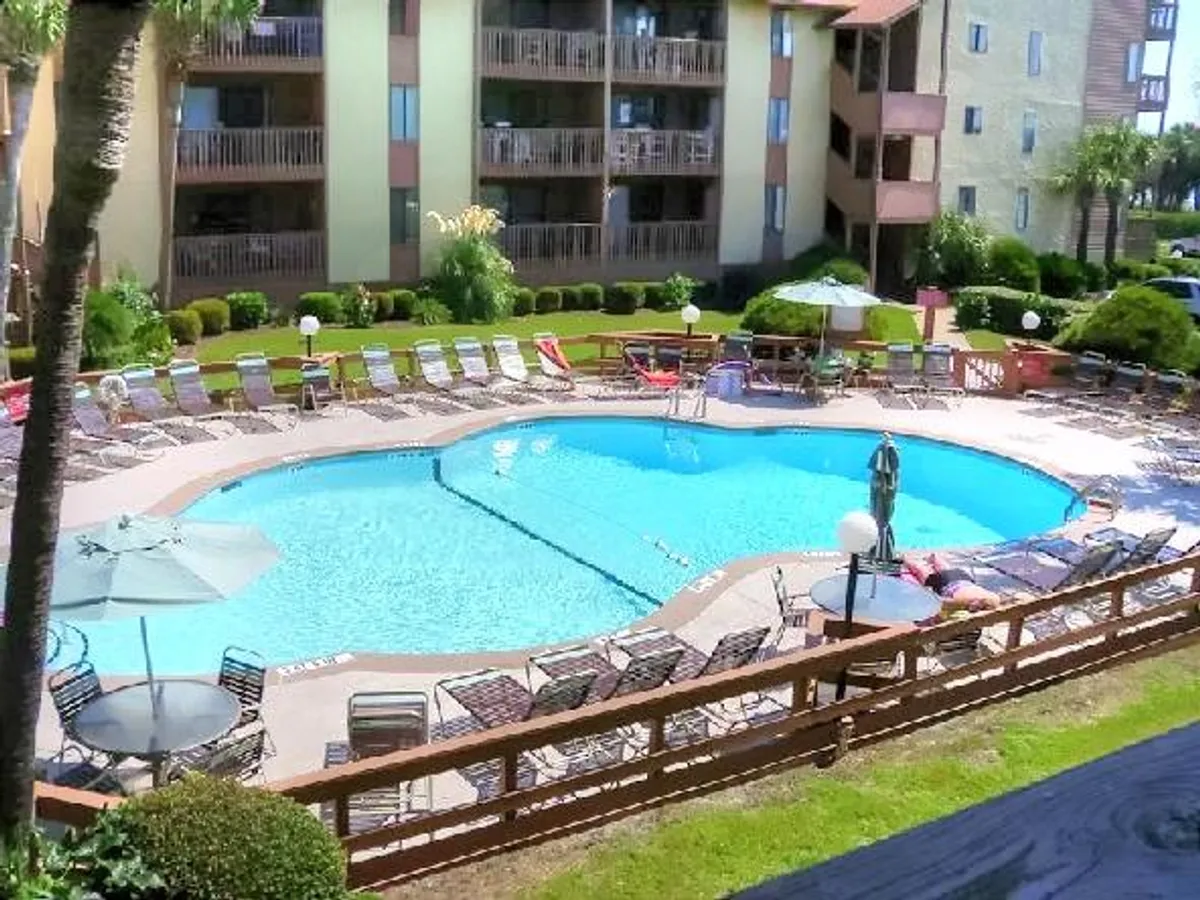 This image shows the resort's inviting outdoor swimming pool surrounded by loungers, landscaped gardens, and the building's balconies. Sunlight bathes the pool area, creating a relaxing, social atmosphere perfect for swimming, sunbathing, and unwinding with friends or family.