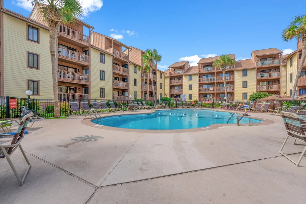 The image showcases the exterior of the Anchorage II condo complex, with a sparkling swimming pool surrounded by lush palm trees and ample sun loungers. The balconies of the apartments overlook the pool area, and the setting sun casts a warm, golden glow over the inviting courtyard, suggesting a relaxing and social atmosphere ideal for both morning swims and sunset gatherings.