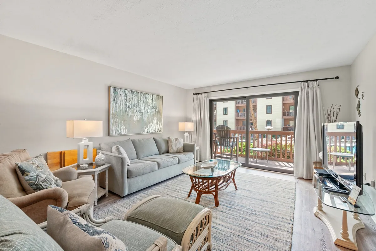 A bright, open living area featuring a comfortable sofa and coffee table set near a large sliding glass door. Warm sunlight pours in, illuminating the cozy, inviting space and drawing attention to artwork on the wall. The view from the glass door hints at a balcony and the ocean beyond, reinforcing the condo’s coastal ambiance.